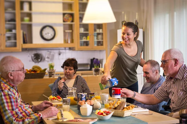Zorgmedewerker schenkt melk in aan een ontbijttafel met bewoners in een woonkeuken